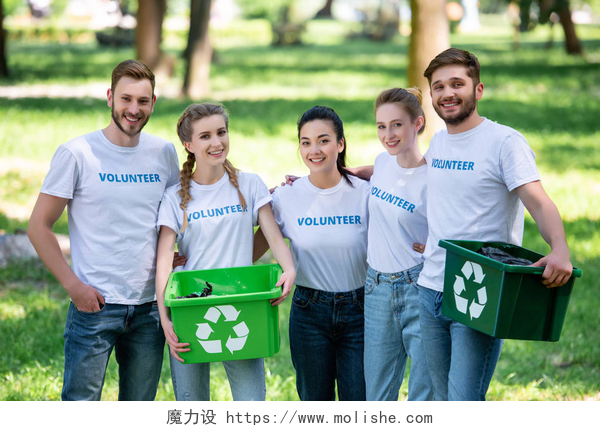 五个年轻的志愿者拿着两个回收箱子在拍照青年志愿者绿色回收箱的垃圾站在公园 